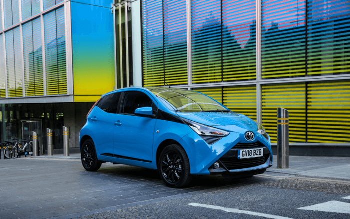 Blue Toyota Aygo hatchback parked in front of a modern building with colourful blue and yellow tinted glass panels. The car has black alloy wheels and a UK registration plate GV18 BZB. The image highlights the compact city car suitable for new cars or used cars market.
