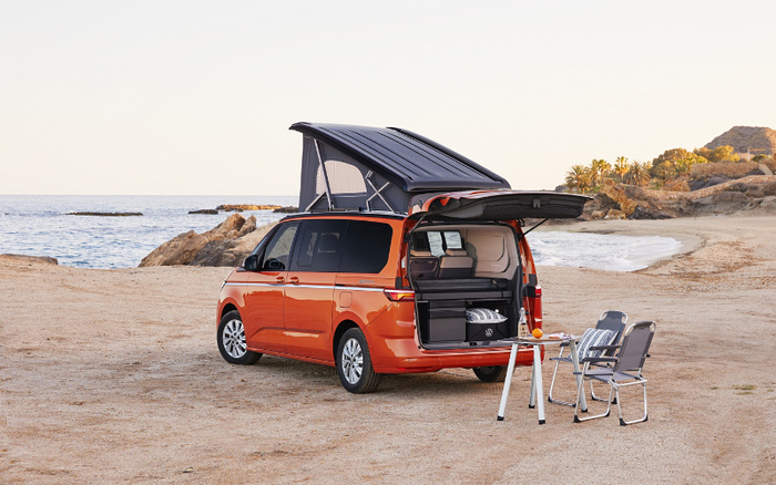 Orange camper van with pop-up roof and open rear door showing interior storage compartments parked on a sandy beach near the sea. Two folding chairs and a small table with drinks on the sand beside the van. Scenic coastal background with rocks and palm trees. Ideal setting for new cars or used cars suitable for camping and outdoor adventures.