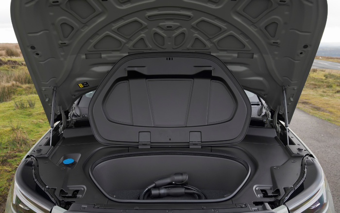 Open front trunk of an electric car showing a charging cable inside the storage compartment, parked on a rural roadside with grass and fields in the background, suitable for used cars and new cars buyers interested in electric vehicles.