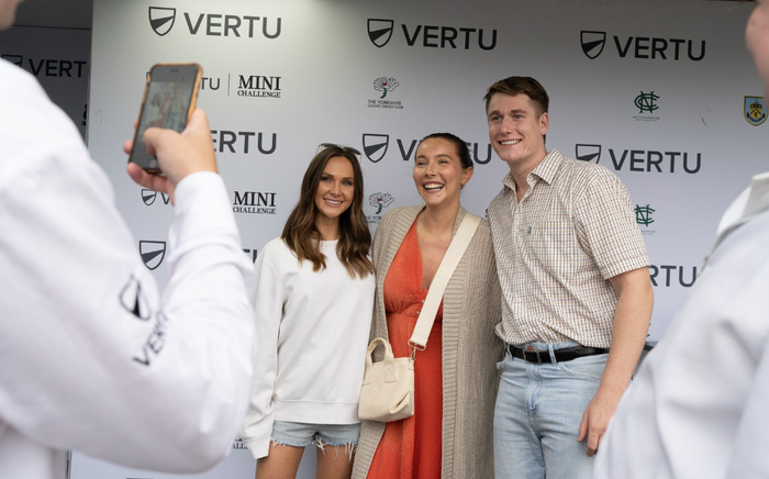 Three people posing for a photo in front of a backdrop featuring the Vertu logo, The Yorkshire County Cricket Club emblem, Nottinghamshire County Cricket Club logo, Burnley FC badge, and text including 'VERTU MINI CHALLENGE'. Two individuals in white shirts with the Vertu logo are taking photographs. The scene captures a casual outdoor event atmosphere.
