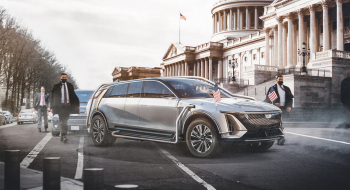 Futuristic silver limousine with Cadillac emblem parked on a street near a government building with large columns and an American flag on top. The limousine has two small American flags on its front, sleek aerodynamic design, and appears to be an electric vehicle with visible charging ports and cables. Several people in suits and face masks are walking nearby, with other cars in the background. This image highlights advanced new cars technology in contrast to used cars on the street.