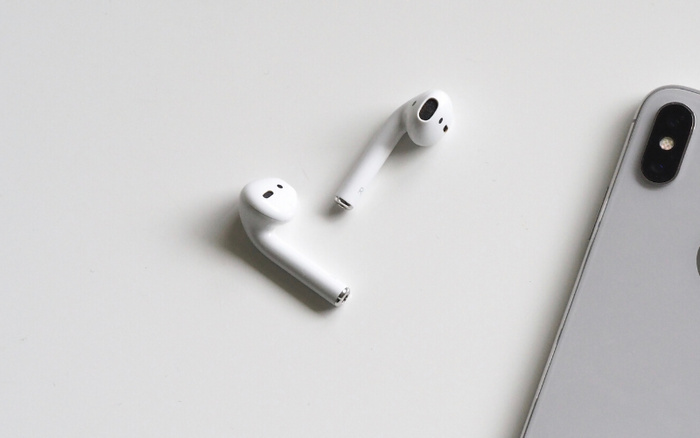 White wireless earbuds placed on a light grey surface next to the top right corner of a silver smartphone showing dual rear cameras, focusing on the sleek design and modern technology without any reference to used cars or new cars.