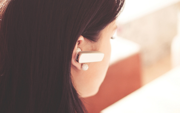 Close-up of a woman wearing a white wireless Bluetooth earpiece and a sparkly round stud earring, with dark brown hair, viewed from the side against a blurred background, symbolising customer service or telephone communication related to used cars and new cars sales.