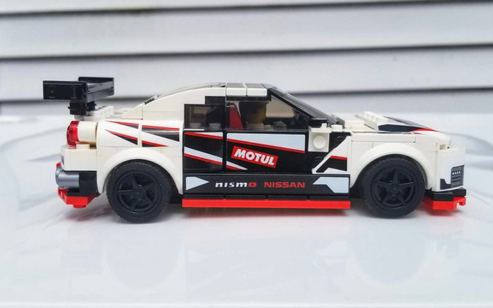 Side view of a detailed Lego model car featuring white, black, and red colours with racing decals. The car displays branding text including 'MOTUL' in red and white on the door, and 'nismo NISSAN' in red and white along the lower side skirt. The model includes a rear spoiler and black wheels with a sporty design, representing a race car style. This image could be used to illustrate used cars or new cars with a focus on race-themed vehicles.