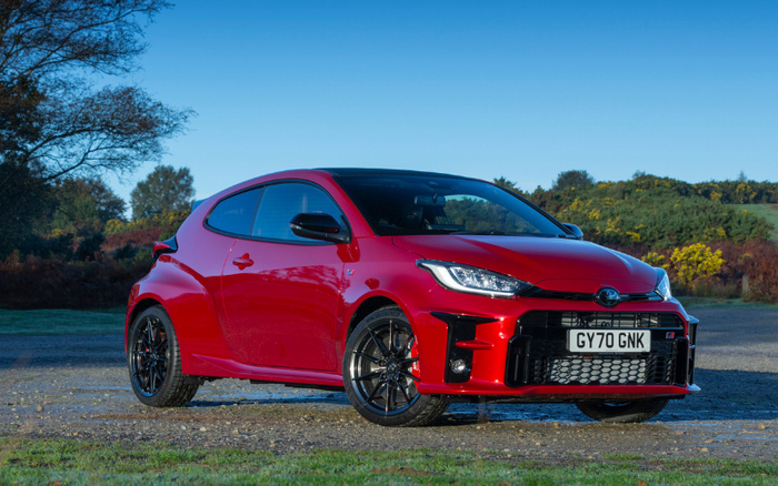 Red Toyota GR Yaris parked outdoors on a gravel surface with greenery and trees in the background under a clear blue sky, showing the front and side profile of this sporty hatchback used car, registration plate GY70 GNK, ideal for buyers interested in used cars and new cars.
