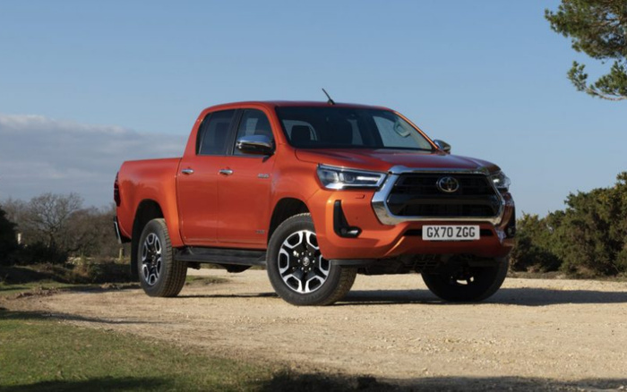 Orange Toyota Hilux pickup truck with UK registration plate GX70 ZGG parked on a dirt road in a rural setting with trees and grass under a partly cloudy blue sky, suitable for showcasing used cars or new cars for sale.