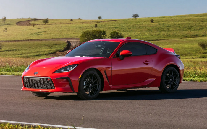 Red sports coupe car parked on a paved road with green rolling hills in the background under clear blue sky, showcasing a sleek modern design with black alloy wheels and tinted windows, suitable for advertisements featuring used cars and new cars.
