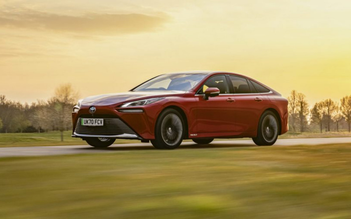 Red Toyota Mirai new car driving on a countryside road at sunset, UK registration plate UK70 FCV, showcasing sleek design and modern headlights, ideal for buyers interested in new cars and used cars.