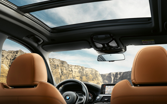 Interior view of a BMW car featuring tan leather seats, a panoramic sunroof, and a dashboard with a navigation screen displaying a map. The car is parked with a scenic rocky cliff landscape visible through the windows. Ideal for showcasing used cars and new cars with advanced features and luxury interiors.