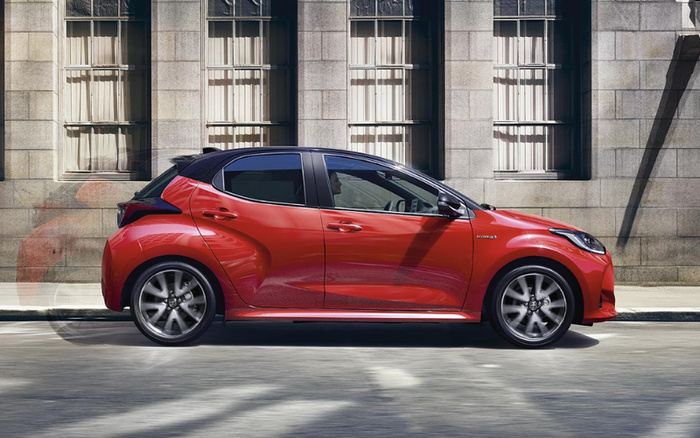 Side view of a red compact hybrid car parked on a city street in front of a stone building with tall windows. The car features sleek, modern styling with a black roof and five-spoke alloy wheels. Keywords included: used cars, new cars.