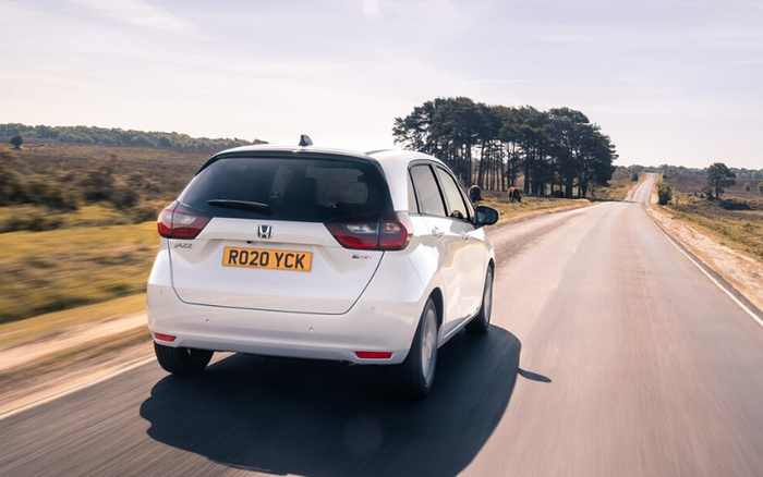 Rear view of a white Honda Jazz RS hybrid car with the registration plate RO20 YCK driving on a countryside road with trees and open fields alongside. Suitable image for used cars and new cars listings.