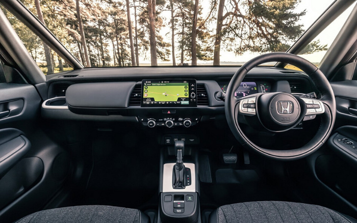 Interior view of a new Honda car showing the dashboard and steering wheel with Honda logo. The dashboard features a central touchscreen display with a navigation map, climate control buttons below, and gear shift in the centre console. The car is parked in a wooded area, visible through the windshield. Suitable for used cars and new cars listings highlighting modern vehicle interiors.