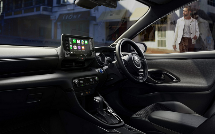Interior view of a modern Toyota car showing the dashboard, steering wheel, and infotainment screen displaying apps like Maps, Phone, and Music. The dashboard features digital climate control set to 21.0 degrees Celsius on the left and 20.5 degrees Celsius on the right. The interior has black leather seats and trim. Outside the car window, a man in a white jacket and brown pants is walking on a city street. This image represents both used cars and new cars with a focus on advanced technology and comfort.