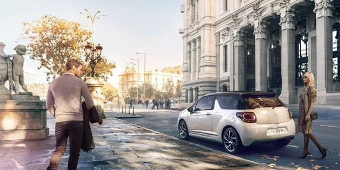 A silver compact car parked on a city street with historic buildings in the background, a man approaching from the left and a woman standing on the right. The scene is set in a bright urban environment with autumnal trees and classic street lamps, ideal for advertising used cars and new cars.
