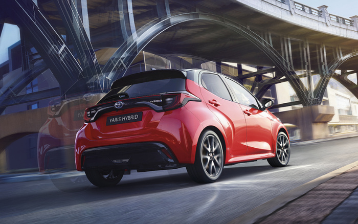 Red Toyota Yaris Hybrid driving on an urban road under a large concrete bridge, showcasing the rear view of the compact hybrid car with the badge 'YARIS HYBRID' visible, ideal for buyers interested in new cars and used cars.