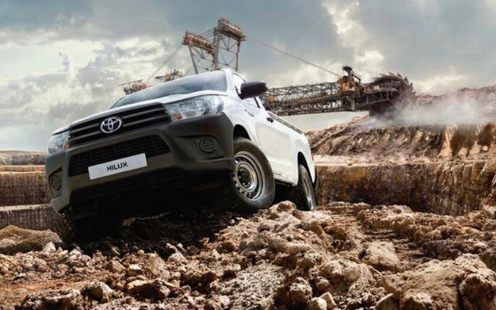 White Toyota Hilux pickup truck navigating rough, muddy terrain at a mining or construction site with large industrial machinery in the background, showcasing the vehicle's off-road capabilities suitable for both used cars and new cars buyers.