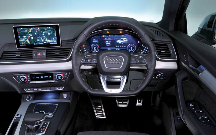 Interior view of a modern Audi car dashboard featuring a digital instrument cluster showing a car graphic, speedometer, and various driving information. The multimedia screen displays a navigation map. The steering wheel is branded with the Audi logo and includes control buttons. The centre console has climate control settings and gear shift lever. This image highlights the advanced technology and design of Audi's used cars and new cars.