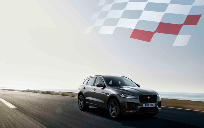 Grey Jaguar SUV driving on an open road near the coast with a large checkered racing flag graphic in the sky. The vehicle has the registration plate 807 YNT. The scene evokes motion and speed, suitable for advertising used cars and new cars.