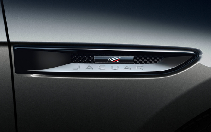 Close-up of the side vent of a grey Jaguar car featuring the Jaguar nameplate and a small emblem with a checkered pattern and red accents, symbolising luxury and performance. Ideal image for used cars and new cars promotions highlighting premium vehicle details.