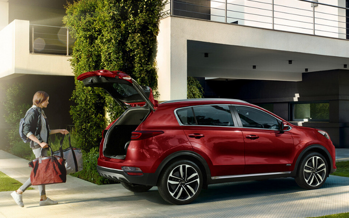 Red Kia Sportage SUV with rear hatch open parked outside a modern house, a woman carrying two large bags walking towards the car, suitable for families looking for used cars or new cars with spacious boot and stylish design.