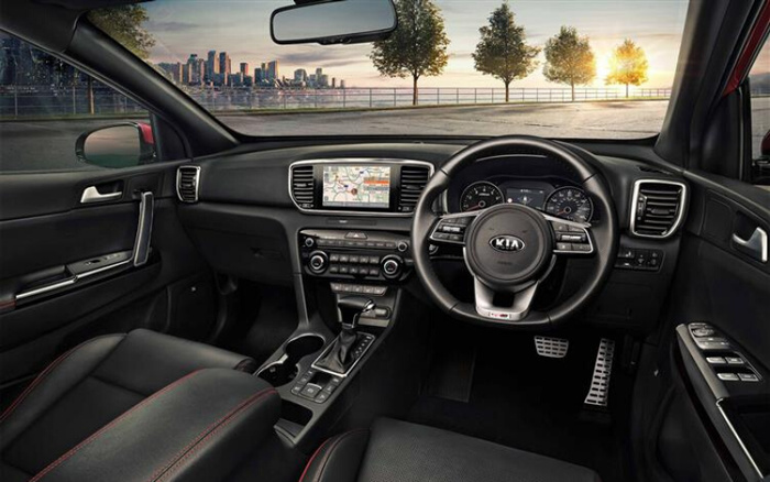 Interior view of a modern Kia car featuring a black leather dashboard and seats with red stitching, a multifunction steering wheel with the Kia logo, a central touchscreen displaying a navigation map, and controls for air conditioning and media. The photo is taken from the passenger seat, showing the driver's side including the gear shift, door controls, and pedals, with a cityscape and trees visible through the windshield. Ideal for showcasing used cars or new cars with advanced features.