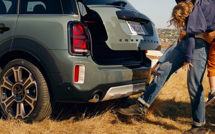 Close-up of the rear of a green Mini Countryman with the boot open, parked on dry grass. A child wearing jeans and a dark top is playfully kicking the back of the car while being held by an adult. The car features distinctive taillights and large alloy wheels. This scene represents a casual outdoor moment with a new or used car.