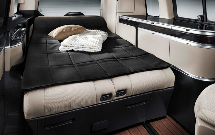 Interior view of a luxury vehicle showcasing a flat, black padded sleeping area with a beige cushion and white striped blanket placed on top. The design features beige upholstery with modern storage compartments and metallic trim, highlighting the spacious and comfortable setup typical in new cars, suitable for rest or travel. This image is relevant for used cars and new cars highlighting versatile vehicle interiors.
