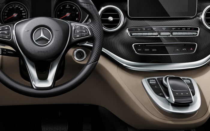 Interior dashboard of a luxury Mercedes-Benz car showing the steering wheel with Mercedes logo, instrument cluster with speedometer and tachometer, central air vents, multimedia control buttons including NAVI, RADIO, MEDIA, TEL, and hazard lights, climate control buttons with AUTO, MENU, A/C, and temperature settings, and a touchpad controller on the beige centre console, highlighting features typical of used cars and new cars in the premium segment.