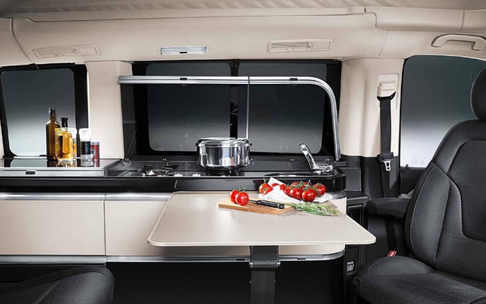 Interior of a modern camper van showing a built-in kitchenette with a stove, sink, and counter space. On the counter are bottles of oil and condiments. A pot sits on the stove, and in front of the kitchenette is a fold-out table with fresh tomatoes, a knife, and herbs on a wooden chopping board. The interior features beige cabinets, black countertop, and black swivel seats, highlighting a compact and functional design suitable for used cars or new cars with camper conversions.