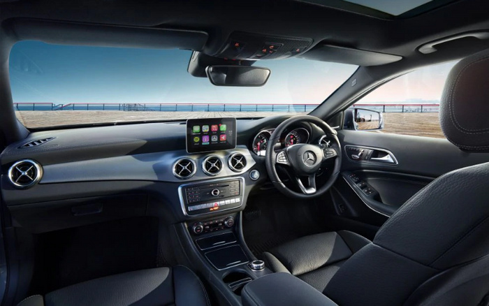 Interior of a modern Mercedes-Benz car showing the driver's side dashboard and steering wheel with the Mercedes logo. The dashboard features a central infotainment screen displaying app icons, three circular air vents, and control buttons below. The car has black leather seats and a panoramic sunroof. View outside shows a clear sky and a waterfront promenade. The image highlights features relevant for buyers considering used cars or new cars.