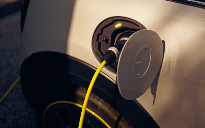 Close-up of an electric vehicle charging port with a yellow charging cable plugged in, showing the round charging socket and open fuel door on a metallic beige or light gold car, highlighting the modern feature often available in new cars and increasingly common in used cars.