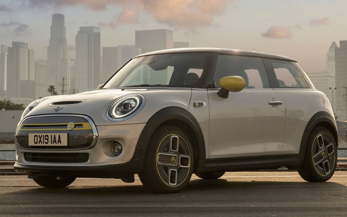Silver Mini Cooper SE electric car with yellow accents on side mirrors, front grille, and wheel rims, parked on urban street with city skyline in the background, UK license plate OX19 IAA, suitable for used cars and new cars.