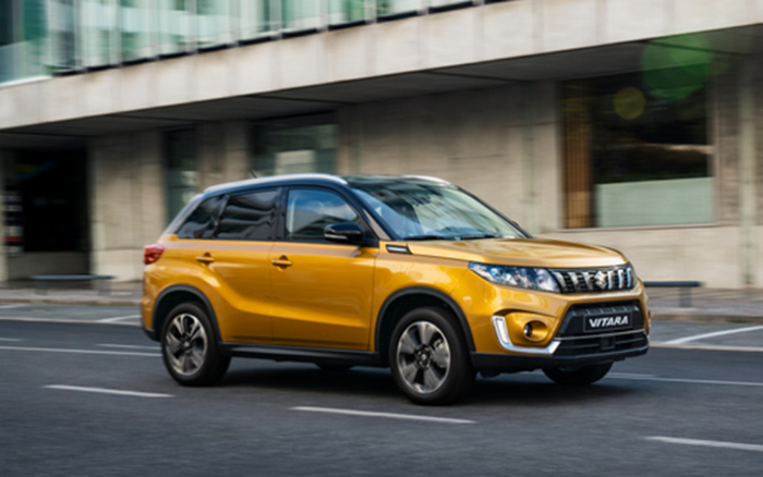 Yellow Suzuki Vitara compact SUV driving on a city street near modern buildings, representing a popular model available as both new cars and used cars.