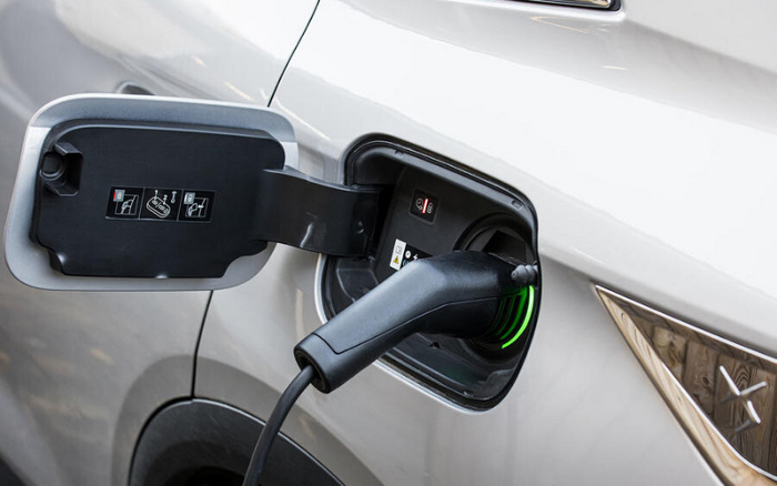 Close-up of an electric car charging port with a charging plug inserted into a white vehicle, showing the charging indicator lights. The inside of the charging port cover has warning and instruction icons. This image relates to used cars and new cars with electric charging capabilities.