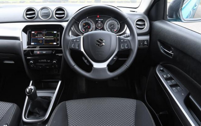 Interior view of a Suzuki car featuring a multifunction steering wheel with the Suzuki logo, a digital dashboard with speedometer and tachometer, a central infotainment system displaying navigation, manual gear stick, and control buttons on the door panel. Suitable for showcasing used cars and new cars interiors.