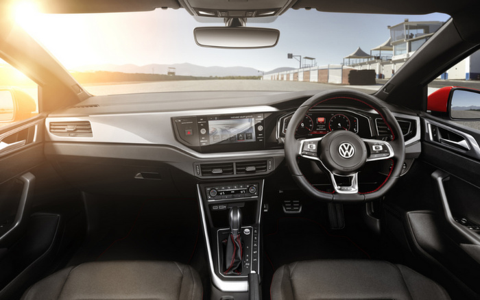 Interior view of a right-hand drive Volkswagen car featuring a modern dashboard with digital display screens showing navigation and car controls, a multifunction steering wheel with the VW logo, automatic gear shift, and comfortable front seats, captured in bright daylight with a clear view of an empty road and distant hills outside. Ideal image representing both used cars and new cars.
