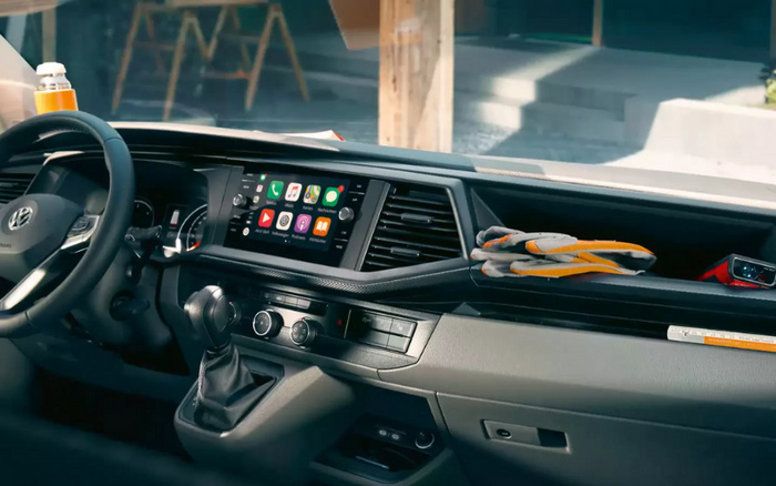 Interior dashboard view of a Volkswagen vehicle showing steering wheel with Volkswagen logo, central touchscreen display with various app icons, gear shift, air conditioning controls, and a pair of work gloves placed on the passenger side dashboard. This modern car interior features digital connectivity and manual gear controls, relevant for new cars and used cars buyers looking for vehicle technology and condition.
