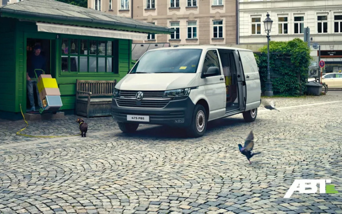 White Volkswagen van with license plate 475 PBS parked on a cobblestone street near a green wooden building. A person is unloading boxes using a trolley. A small dog and pigeons are on the street. ABT logo is visible in the bottom right corner. Image relates to used cars and new cars, focusing on commercial vehicle transport.