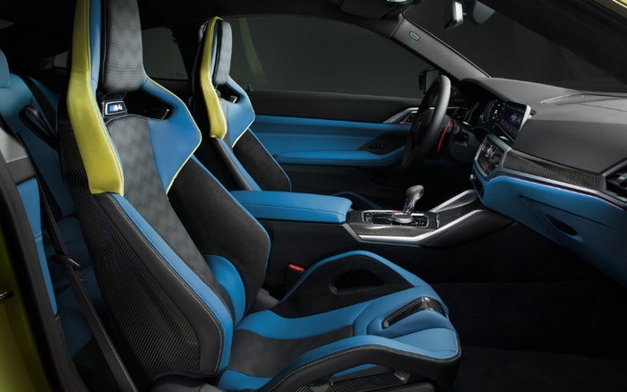 Interior of a BMW M4 sports car showcasing blue and black leather and suede sports seats with yellow accents and M4 badges, carbon fibre trim on the seats and dashboard, modern centre console with gear selector, digital driver display, and blue door panels, highlighting features relevant for buyers interested in premium used cars or new cars.