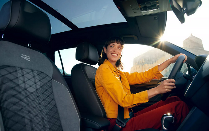 Young woman smiling and driving a modern car with a panoramic sunroof, comfortable interior featuring black and grey seats with an 'R' logo, in bright sunlight with architectural buildings visible outside, showcasing the enjoyment of driving new cars and suitable for used cars buyers.