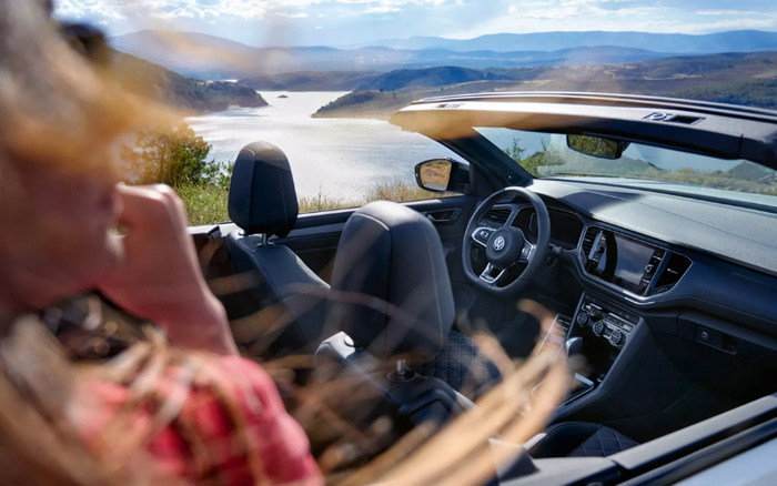 Interior view of a Volkswagen convertible car with black leather seats and modern dashboard, parked with the roof down overlooking a scenic lake and mountain landscape. A woman with blonde hair wearing a red plaid shirt is sitting in the passenger seat, gazing at the view. The image highlights comfort and style, ideal for advertising used cars and new cars.