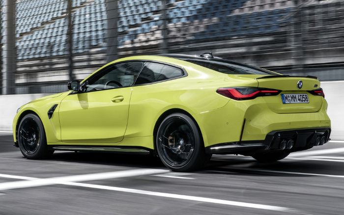 Yellow BMW M4 coupe driving on a city street with modern buildings in the background, showcasing a sleek and sporty design with black alloy wheels and quad exhaust pipes, ideal for advertising used cars and new cars.