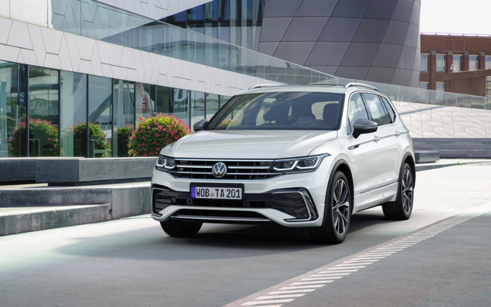 White Volkswagen Tiguan SUV parked in front of a modern glass building with reflections of plants, showing the front and side view. The car has a German license plate reading 'WOB TA 201'. Ideal for used cars or new cars search.