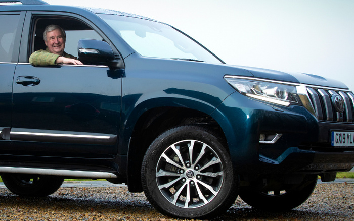 Smiling elderly man leaning out of the driver's side window of a dark blue Toyota SUV parked on a gravel surface, showcasing a polished alloy wheel and modern headlight design, with a visible UK license plate GX19 YL indicating a new car. The image represents both used cars and new cars options.